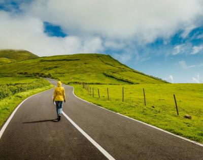 A woman walking down a winding road with an open field and clouds in the blue sky