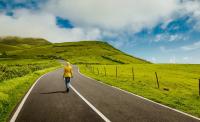 A woman walking down a winding road with an open field and clouds in the blue sky